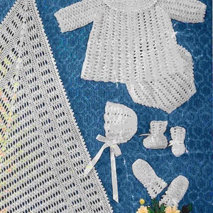 Op de afbeelding: Een witte gehaakte babyset inclusief een deken, een muts, slofjes en wanten. De set is gelegd op een blauwe achtergrond.