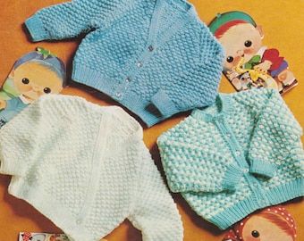May include: Three knitted baby cardigans in light blue, white, and teal. The cardigans feature a textured pattern and button fastenings. Children's book illustrations are scattered around the sweaters, adding a playful touch to the scene.