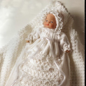 May include: A white knitted baby doll dress with lace trim and a satin ribbon. The doll is wearing a white knitted bonnet with lace trim.