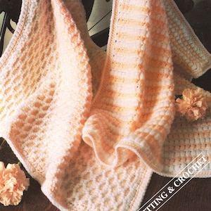 May include: A soft, hand-knitted blanket in shades of peach and cream. The blanket features a textured pattern with a border and is draped over a dark surface. The words "Knitting & Crochet" are visible in the lower right corner.
