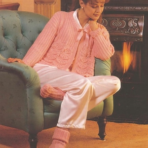 Peut inclure: Une femme portant un cardigan en tricot rose avec un motif festonné est assise dans un fauteuil vert devant une cheminée. Elle porte un pantalon blanc en satin et des chaussettes roses.