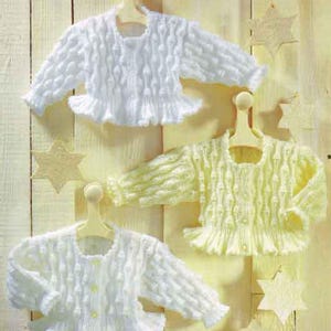 Puede incluir: Tres cárdigans de bebé de punto en blanco y amarillo pálido, expuestos en perchas de madera. Los suéteres tienen mangas largas, cierres de botones y dobladillos con volantes. Fondo de panel de madera clara con estrellas.