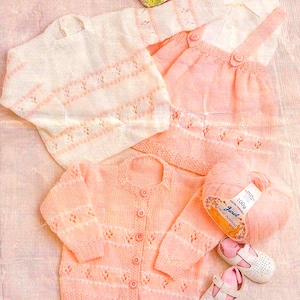 Pode incluir: Um conjunto de roupas de bebé de malha em rosa e branco. O conjunto inclui um casaco de malha, um vestido e uma bola de lã. A lã está etiquetada como "Jarlo New Aral 100g".