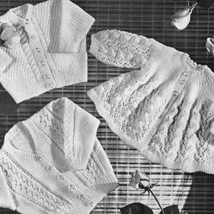 Puede incluir: Tres jerséis de punto blanco para bebé con diferentes patrones. Los jerséis están colocados sobre una superficie tejida con rosas.