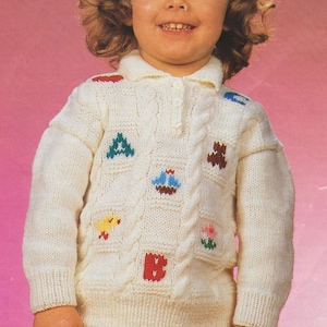 Puede incluir: Un suéter de punto color crema con cuello y mangas largas. El suéter presenta detalles de punto trenzado y parches bordados coloridos de letras, pájaros y flores. El niño lleva pantalones blancos.