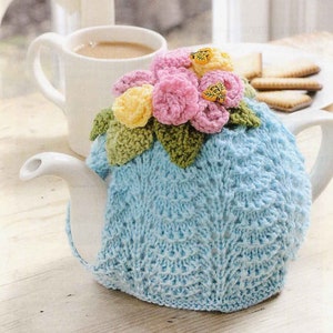 Puede incluir: Un calienta-tetera de punto azul con un diseño de flores de ganchillo en rosa, amarillo y verde. El calienta-tetera está en una tetera blanca.