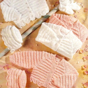 Op de afbeelding: Twee sets gebreide babykleding, een in wit en een in roze. Elke set bevat een trui, een muts en slofjes. De truien hebben een knoopsluiting en een gegolfde rand. De mutsen zijn simpel en de slofjes hebben een gegolfde rand.