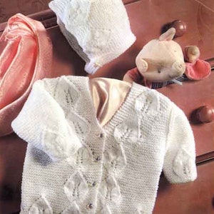 May include: A white knitted baby cardigan with leaf-shaped details and matching bonnet. The cardigan has a V-neck and button closure. The bonnet has a brim and leaf-shaped details. The set is made of a soft, textured yarn.