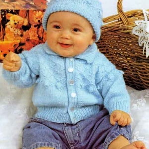 Può includere: Un bambino che indossa un cardigan di lana blu chiaro con chiusura a bottoni e un cappello di lana blu chiaro abbinato. Il bambino è seduto su una superficie bianca e indossa jeans blu.