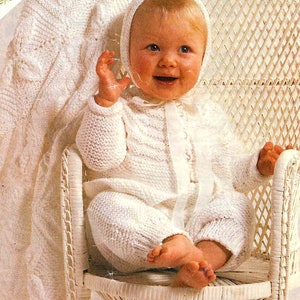 May include: A white knitted baby outfit with a matching bonnet. The outfit features a long-sleeved sweater and pants with a button closure. The baby is sitting in a white wicker chair.