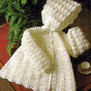 May include: A white, crocheted baby outfit, including a hooded jacket and matching bonnet. The jacket has a scalloped hem and is fastened with three buttons. The set is made with a textured stitch pattern, creating a puffy, three-dimensional effect.