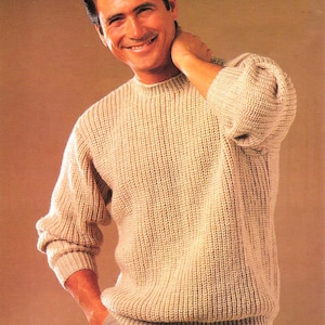 Peut inclure: Un pull beige en maille côtelée avec un col rond et des manches longues. Le pull a une coupe décontractée et est porté avec un pantalon gris. L'image montre une personne souriante portant le pull.