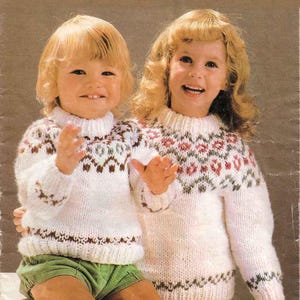 May include: Two children wearing matching white knit sweaters with patterned yoke designs in brown, red, and green. The sweaters have ribbed collars and cuffs. One child wears green shorts.