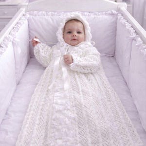 Può includere: Un abito da battesimo bianco a maniche lunghe con un intricato motivo a maglia, una cuffietta abbinata con bordo arricciato e un nastro di raso verticale centrale. Il capo è realizzato in morbido filato bianco con delicati dettagli in pizzo.