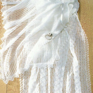 May include: White crocheted baby blanket with a delicate lace pattern. The blanket is folded and draped over a wooden surface.