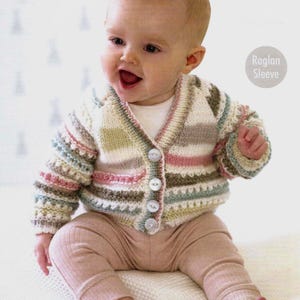 以下が含まれることがあります： ボタン留めのストライプカーディガンを着た赤ちゃん。カーディガンはラグランスリーブで、ピンク、グリーン、ホワイトなど様々な色で編まれています。赤ちゃんは白い毛布に座って笑顔です。