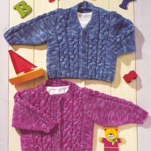 Könnte beinhalten: Zwei Baby-Strickjacken mit Zopfmuster in Blau und Magenta. Die blaue Strickjacke hat einen V-Ausschnitt und die magenta eine Rundhalsausschnitt. Holzspielzeug-Segelboot, Auto und Buchstaben mit der Aufschrift "BABY" sind ebenfalls im Bild.