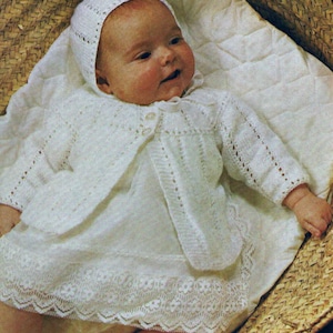 Könnte beinhalten: Ein weißes gestricktes Baby-Outfit mit passender Mütze. Das Outfit hat einen Spitzenbesatz und einen Knopfverschluss. Das Baby liegt in einem Weidenkorb.