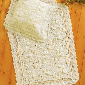 May include: Cream-coloured, knitted baby blanket and pillow set. The blanket features a repeating floral pattern within diamond shapes, with a scalloped edge. The pillow has a matching floral design. The set is placed on a wooden surface.