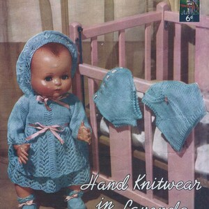 May include: A blue knitted doll outfit with a matching bonnet, booties, and a pink ribbon. The doll is wearing the outfit and is standing next to a pink crib. The text "Hand Knitwear in Lavenda" is printed on the image.