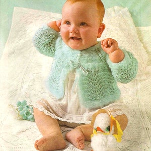 May include: A baby wearing a light green knitted cardigan with a white lace trim and a white dress. The baby is sitting on a white crocheted blanket and holding a white stuffed duck with yellow accents.