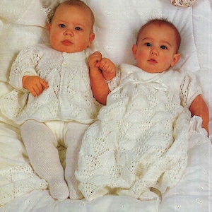 May include: Two babies wearing white crocheted outfits. The baby on the left is wearing a cardigan and pants, while the baby on the right is wearing a dress. Both babies are lying on a white blanket.