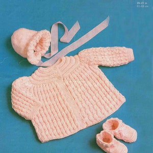 May include: A light pink knitted baby set, including a bonnet, sweater, and booties. The sweater and booties have a ribbed design. The bonnet has a ruffled edge and a ribbon. The text "Baby D K" is visible in the upper right corner. The sweater is designed for a baby with a chest size of 51-53 cm.