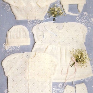 May include: A set of white knitted baby clothes, including a cardigan, dress, onesie, hat, and socks. The clothes are made with a delicate lace pattern.