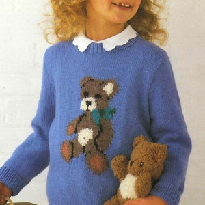 May include: A young girl wearing a blue knitted sweater with a brown teddy bear design. She is holding a brown teddy bear in her right hand.