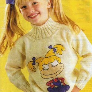 Könnte beinhalten: Ein junges Mädchen trägt einen weißen Strickpullover mit einer Comicfigur auf der Vorderseite. Die Figur ist ein Mädchen mit blonden Haaren und einem lila Kleid. Das Mädchen trägt eine blaue Jeans und hat ihre blonden Haare in Zöpfen mit blauen Schleifen.