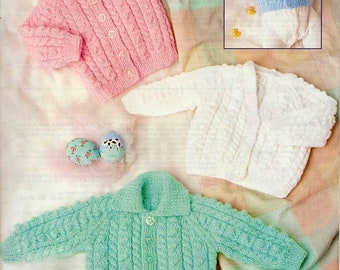 May include: Three knitted baby cardigans in pink, white, and green with a cable knit pattern. The cardigans are laid out on a white and pink blanket.