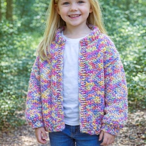 Puede incluir: Una niña con un cárdigan de punto grueso y colorido, con tonos rosas, amarillos y azules. El suéter abierto se combina con una camisa blanca y vaqueros azules. El fondo es un bosque verde borroso.