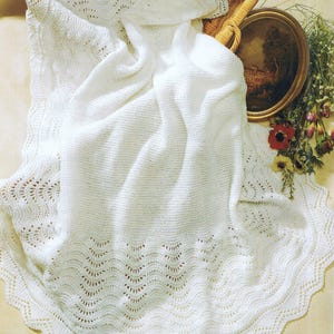 May include: A white knitted blanket with a scalloped edge and a delicate openwork design. The blanket is draped over a wicker basket, with a painting and flowers in the background. The blanket's texture is soft and inviting.