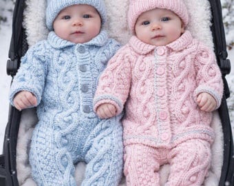 May include: Two infants wearing matching cable-knit outfits in light blue and soft pink. Each outfit includes a long-sleeved button-up romper with integrated booties and a matching beanie topped with a multi-colored pom-pom, resting in a stroller.