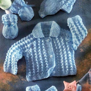 May include: A light blue knitted baby clothing set, including a jacket, hat, mittens, and booties. The set features a textured pattern and is tied with blue ribbons. The jacket has three buttons. A pink star-shaped plush toy is also present.