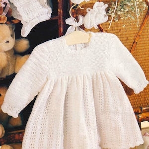 Puede incluir: Vestido de bebé blanco de ganchillo con mangas largas, exhibido en una percha de madera. También se ven un gorro y patucos de ganchillo a juego. El vestido presenta una falda calada detallada y un cuello redondo. La palabra "CROCHET" es visible en la esquina superior derecha.