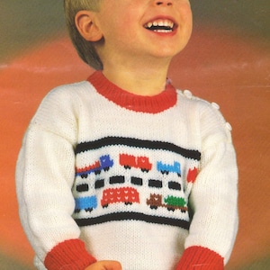 Puede incluir: Un suéter de punto blanco con ribetes rojos y un diseño de coches coloridos en la parte delantera. El suéter tiene puños rojos y un cuello rojo.