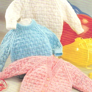 Puede incluir: Tres jerséis de punto para bebé en blanco, azul claro y rosa. Los jerséis tienen un patrón de punto texturizado y se muestran sobre un fondo blanco.