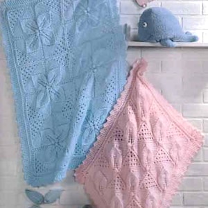 May include: Two crocheted baby blankets, one light blue and one pink, with a delicate floral pattern. The blankets are hanging on a white brick wall with a small stuffed whale and leaves on a shelf above.