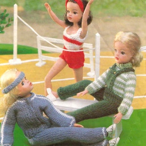 Könnte beinhalten: Drei Puppen in gestrickter Sportkleidung posieren auf einem Miniatur-Leichtathletik-Set. Eine Puppe trägt ein rot-weißes Outfit, die Arme erhoben. Eine weitere Puppe trägt einen blauen Trainingsanzug, und die dritte Puppe grüne Latzhosen und einen gestreiften Pullover.