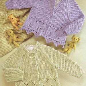 Könnte beinhalten: Zwei gestrickte Baby-Cardigans mit Knöpfen. Ein Cardigan ist lavendelfarben mit einer zarten Spitzenborte und der andere ist hellgrün mit einer ähnlichen Spitzenborte.