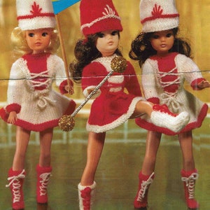May include: Three dolls wearing red and white knitted outfits with matching hats. The dolls are holding a baton, a flag, and a drum.