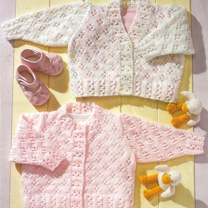 May include: Two knitted baby cardigans, one white and one pink, with button closures. Pink baby shoes and two plush duck toys are also present. The cardigans have a delicate, openwork design, suggesting a handmade or vintage style.