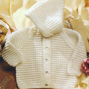 May include: Cream-colored knitted baby cardigan and matching bonnet. The set features a textured stitch pattern, long sleeves, and button closures. The bonnet has a rounded shape, and the cardigan has a classic design.