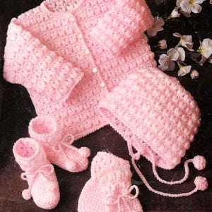 May include: A handmade, light pink crochet baby set, including a button-up cardigan, a bonnet with ties, and matching booties and mittens. The set is made with a textured stitch and features delicate details.