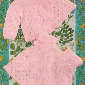 Op de afbeelding: Een lichtroze gebreide baby-cardigan en jurk set. De cardigan heeft lange mouwen en knoopsluitingen. De jurk heeft korte mouwen. Beide items hebben een textuur, openwerk ontwerp. De set wordt gepresenteerd op een stof met bloemenpatroon.