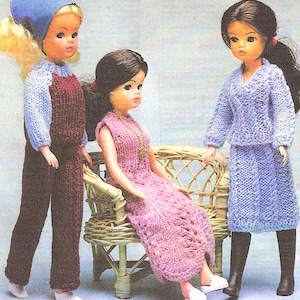 May include: Three dolls wearing knitted outfits. The doll on the left is wearing a blue headband, a light blue sweater, and maroon overalls. The doll in the middle is wearing a pink dress with a gold necklace. The doll on the right is wearing a light purple sweater and skirt.