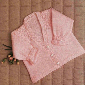 May include: A pink knitted baby cardigan with a V-neck and button closure. The cardigan has a cable knit pattern on the front and sleeves.