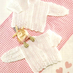 Op de afbeelding: Twee witte, gebreide babyvestjes met knoopsluiting worden getoond op een roze en witte geruite ondergrond. De vestjes hebben een delicaat, opengewerkt ontwerp. Een kleine pop met een gele jurk en gestreepte benen ligt ernaast.