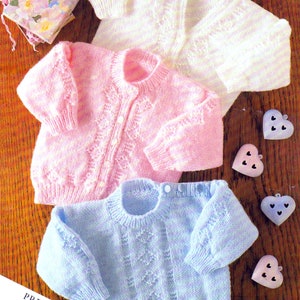 May include: Three knitted baby sweaters in pink, white, and blue with a cable stitch pattern. The sweaters are laid out on a wooden surface with decorative hearts.